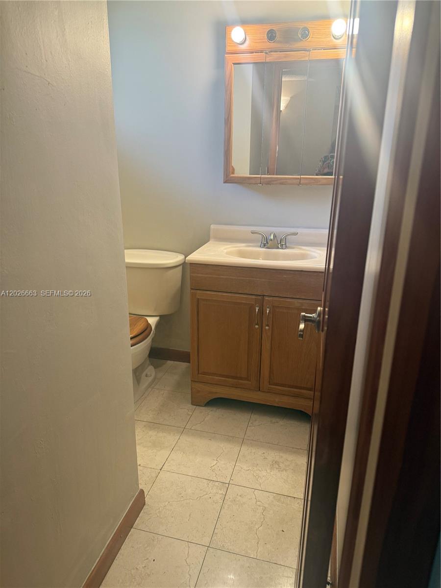 A small bathroom features a wood vanity, a mirror with lights, and a toilet — 100 Ashbury Rd # 104, Hollywood FL 33024, HOLLYWOOD, FL