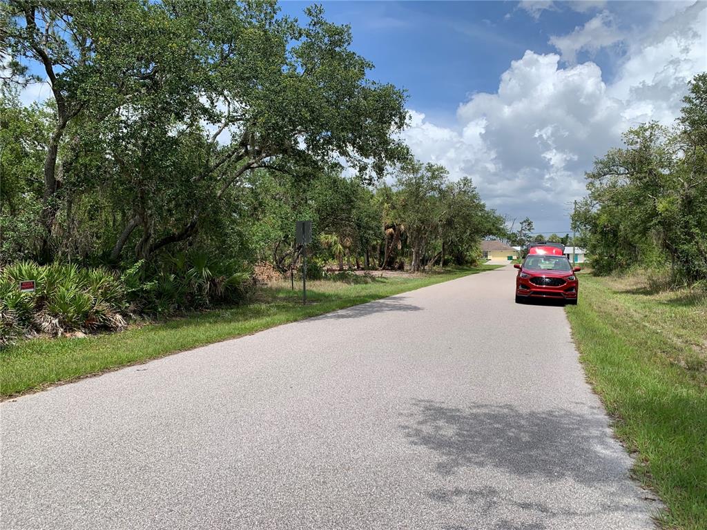 A red SUV is parked on a paved road next to a wooded, undeveloped lot — 17496 BILLIAR AVENUE, PORT CHARLOTTE, FL