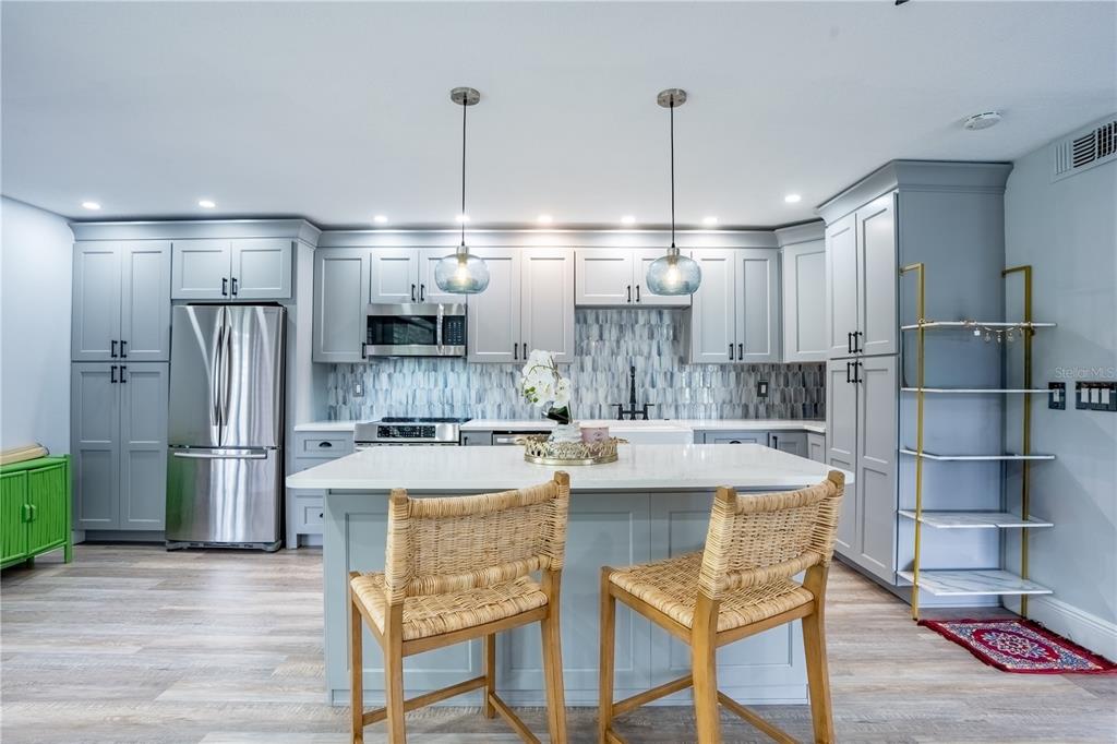 A kitchen features light gray cabinets, a central island with two chairs, and stainless appliances — 1005 S BAYSHORE BOULEVARD UNIT 102, SAFETY HARBOR, FL