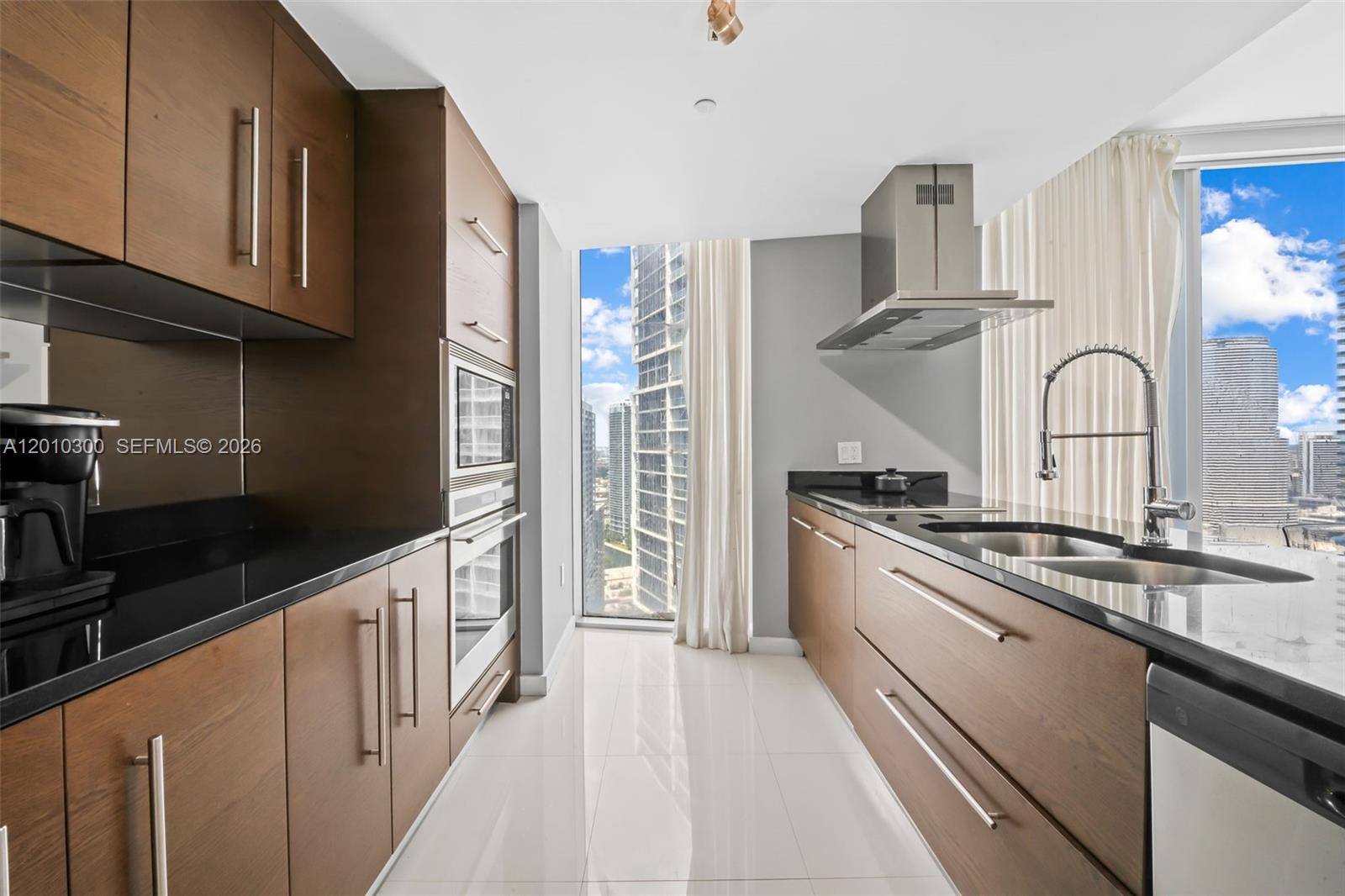 A modern galley kitchen features wood cabinets, black countertops, and floor-to-ceiling city views — 495 Brickell Ave # 3710, Miami FL 33131, MIAMI, FL