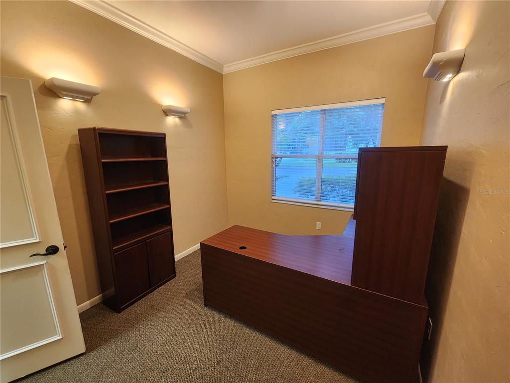 A home office features a wooden desk, a matching bookshelf, beige walls, and carpeted flooring — 101 NW 75TH STREET UNIT Suite 1-A, GAINESVILLE, FL