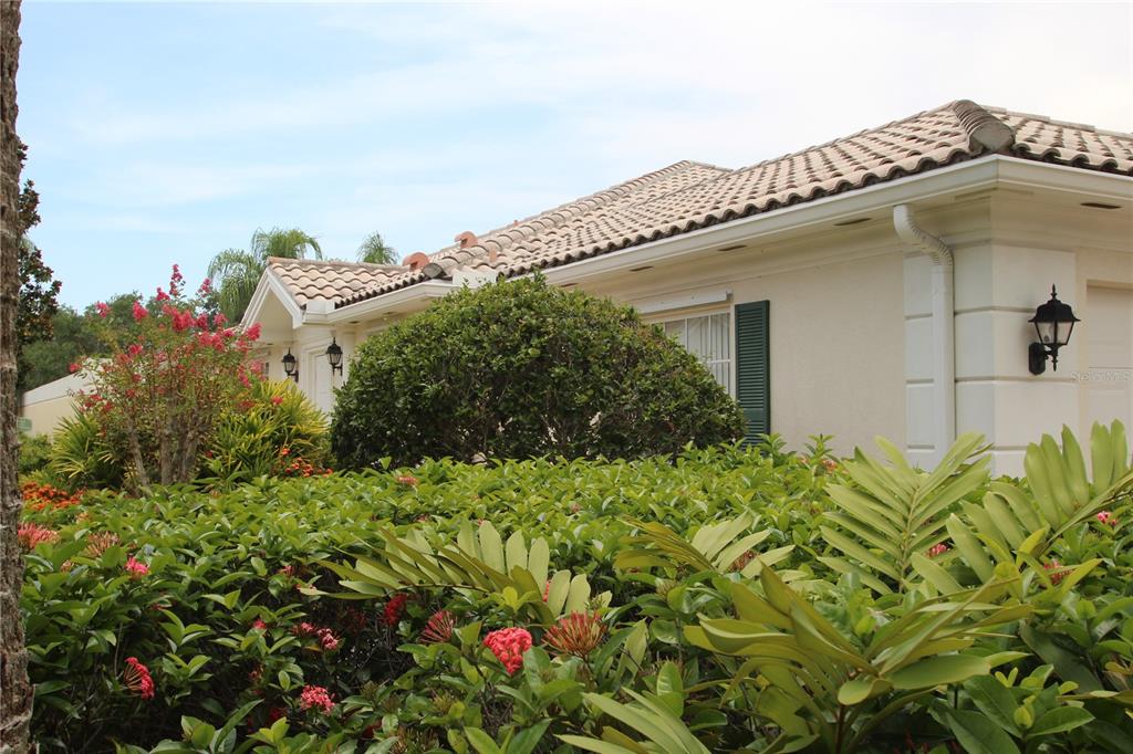 A single-story house with a tiled roof is partially obscured by lush green landscaping — 5506 MODENA PLACE, SARASOTA, FL
