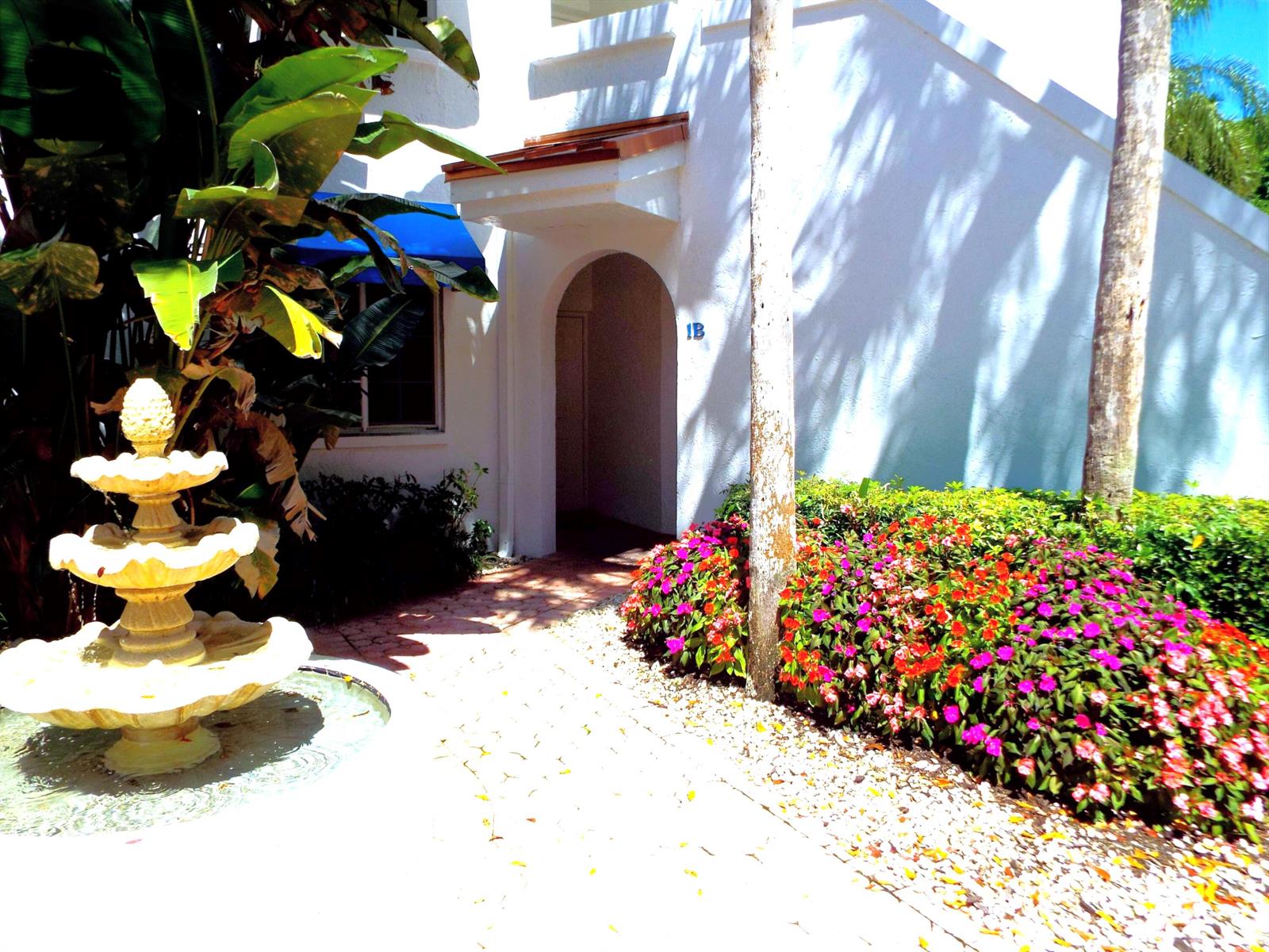 A white building entrance with a fountain, tropical plants, and colorful flowers in the foreground — 4128 W Palm Aire Drive # 281b, Pompano Beach FL 33069, POMPANO BEACH, FL