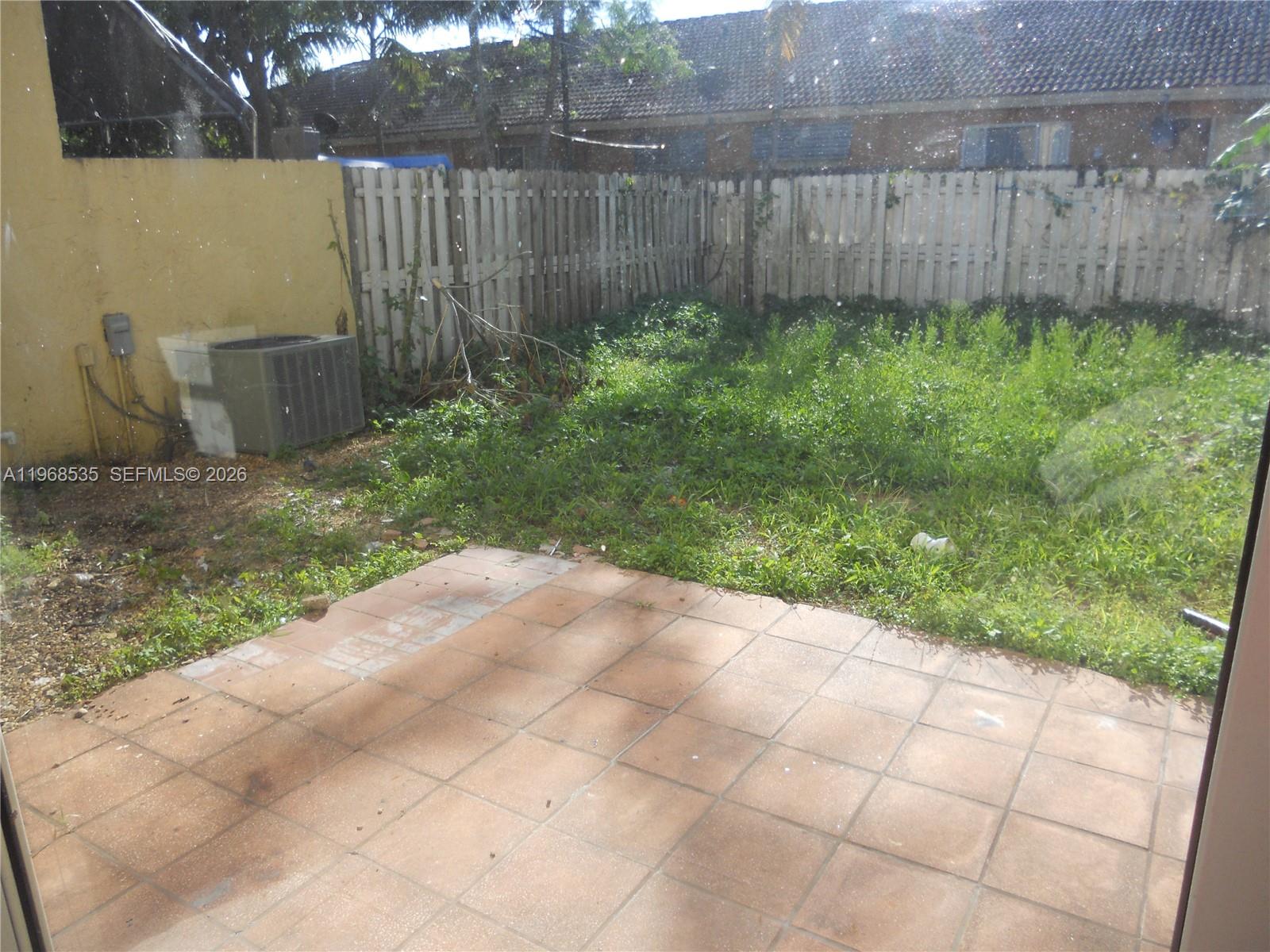 A backyard features a tiled patio, an air conditioning unit, and an overgrown grassy area — 22 SW 14th Ter, Homestead FL 33030, HOMESTEAD, FL