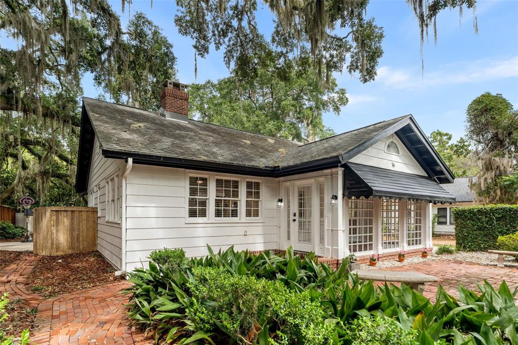 A white cottage with a brick chimney and a brick patio under large oak trees — 16 NW 20TH TERRACE, GAINESVILLE, FL