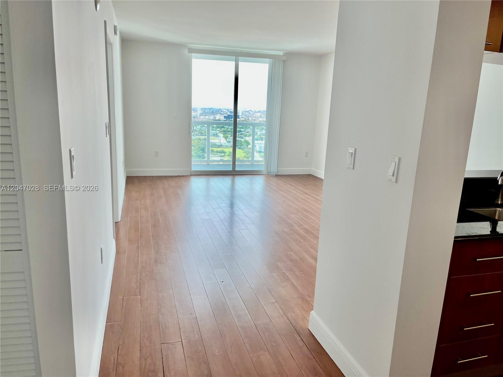 An empty living room features light wood flooring, white walls, and a sliding glass balcony door — MIAMI, FL