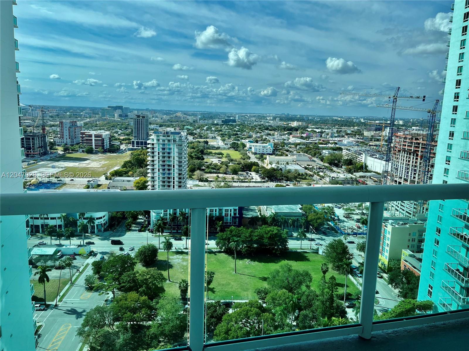 A high-angle view from a balcony overlooking a city with buildings, trees, and construction cranes — MIAMI, FL