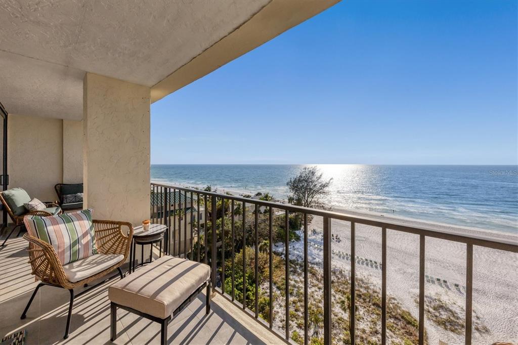 A balcony with wicker chairs and an ottoman overlooks a sandy beach and the ocean — 20 WHISPERING SANDS DRIVE UNIT 903, SARASOTA, FL