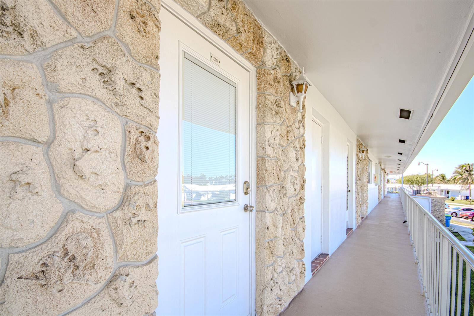 An exterior walkway features white doors with stone accents and a metal railing — 2850 W Golf Boulevard # 210, Pompano Beach FL 33064, POMPANO BEACH, FL