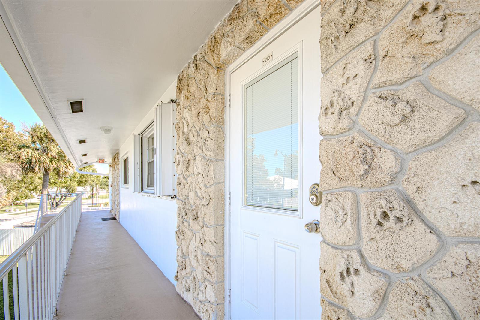 An exterior walkway leads to a white door with a keypad lock and stone facade — 2850 W Golf Boulevard # 210, Pompano Beach FL 33064, POMPANO BEACH, FL