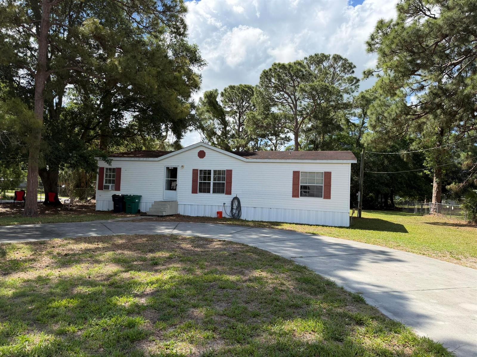 A white single-wide mobile home with red shutters sits on a lot with mature trees — 5924 NE 4th Lane, Okeechobee FL 34974, OKEECHOBEE, FL
