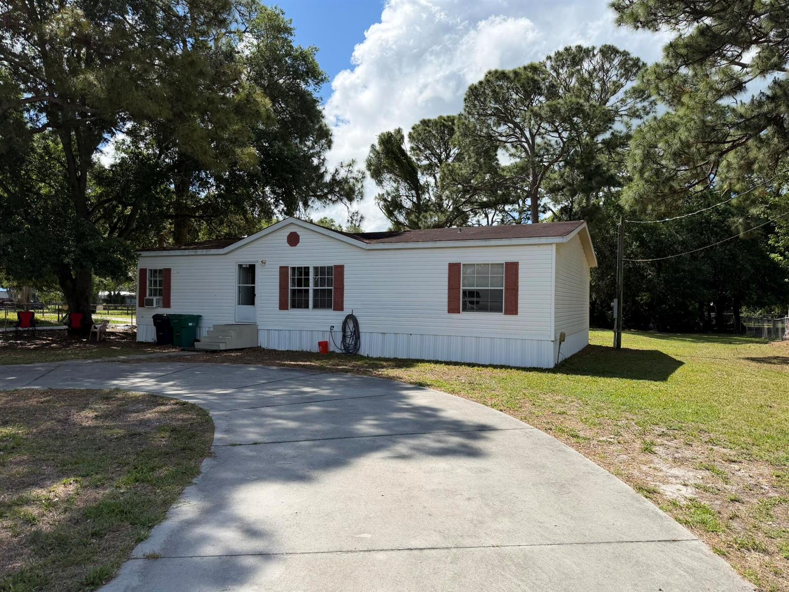 A single-story mobile home with white siding and red shutters sits behind a concrete driveway — 5924 NE 4th Lane, Okeechobee FL 34974, OKEECHOBEE, FL