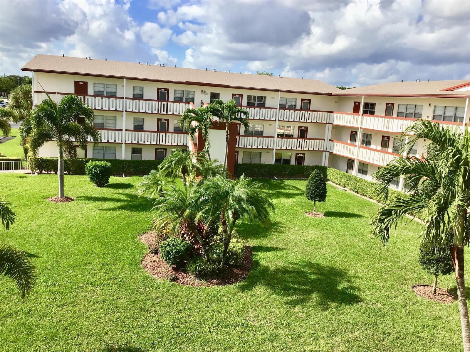A three-story apartment building overlooks a grassy courtyard with palm trees and manicured shrubs — 308 Brighton H # 308, Boca Raton FL 33434, BOCA RATON, FL