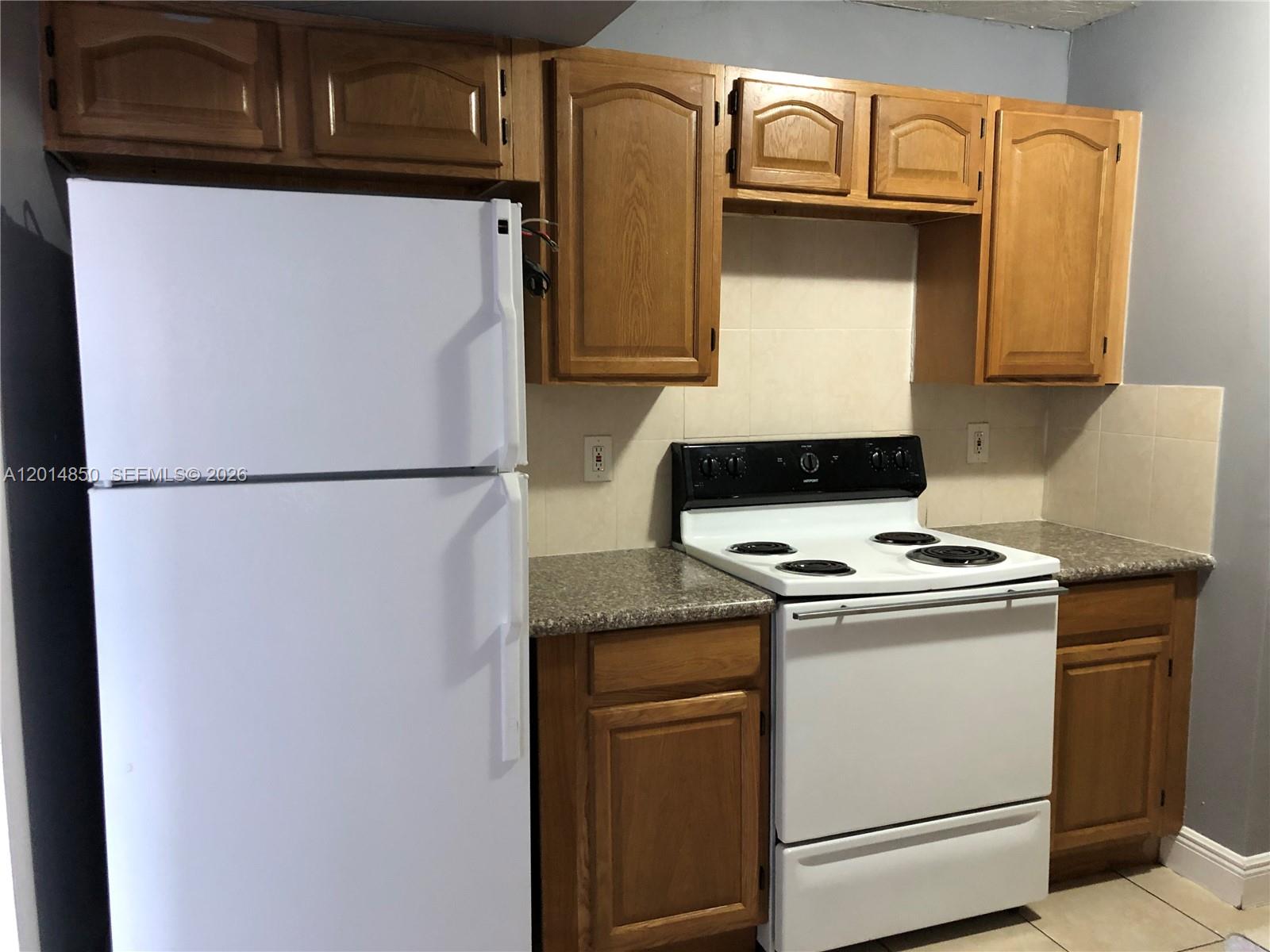 A kitchen features white appliances, wood cabinets, and granite-look countertops against a tiled backsplash — 424 SW 12th Ave, Homestead FL 33030, HOMESTEAD, FL