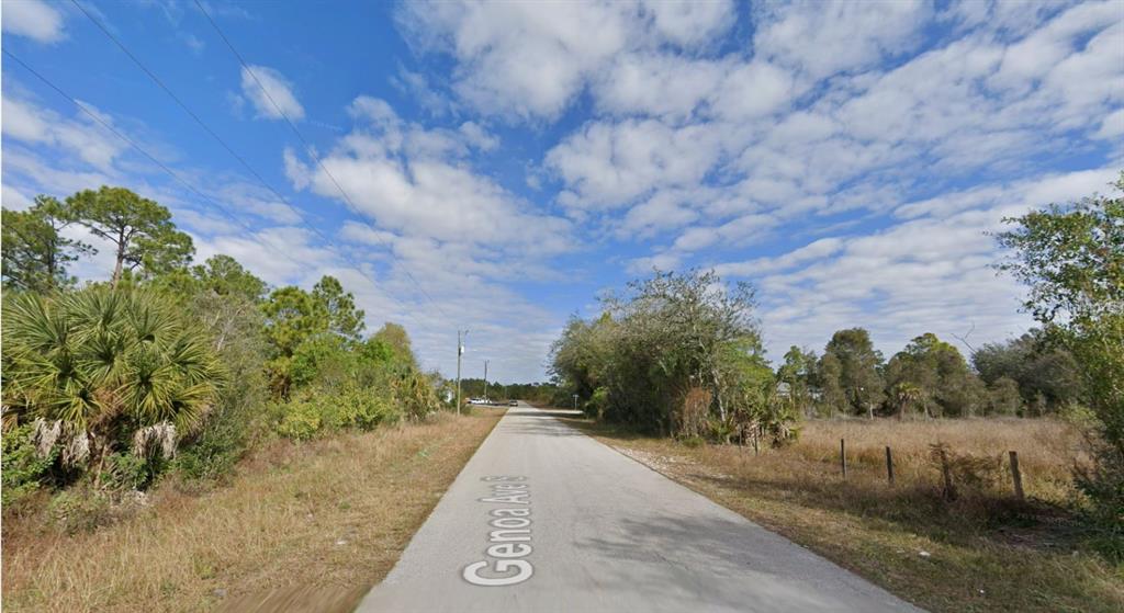A paved road stretches through a rural, wooded area under a blue, cloudy sky — 329 GENOA AVENUE S, LEHIGH ACRES, FL