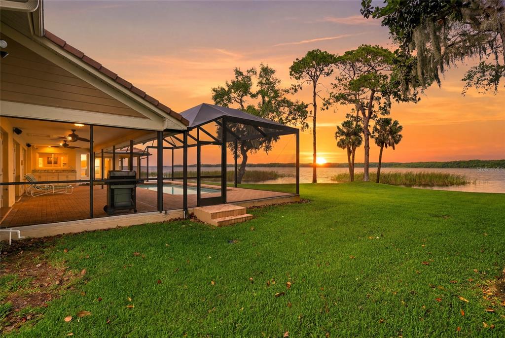 A screened-in pool patio overlooks a lake at sunset with a grassy lawn in front — 3493 SHORELINE CIRCLE, PALM HARBOR, FL