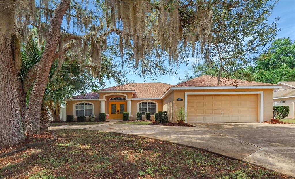 A single-story house with a tan exterior, a two-car garage, and a large oak tree — 34224 SHADEWOOD CIRCLE, LEESBURG, FL