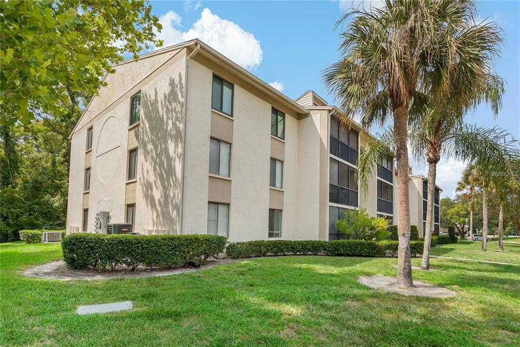 A three-story apartment building with beige stucco walls and screened balconies sits on a lawn — 3161 LAKE PINE WAY S UNIT F3, TARPON SPRINGS, FL
