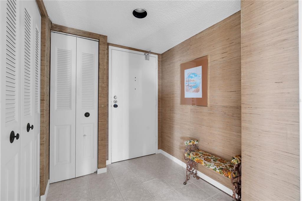 An entryway features light-colored tile flooring, textured wallpaper, white louvered closet doors, and a bench — 7600 BAYSHORE DRIVE UNIT 701, TREASURE ISLAND, FL