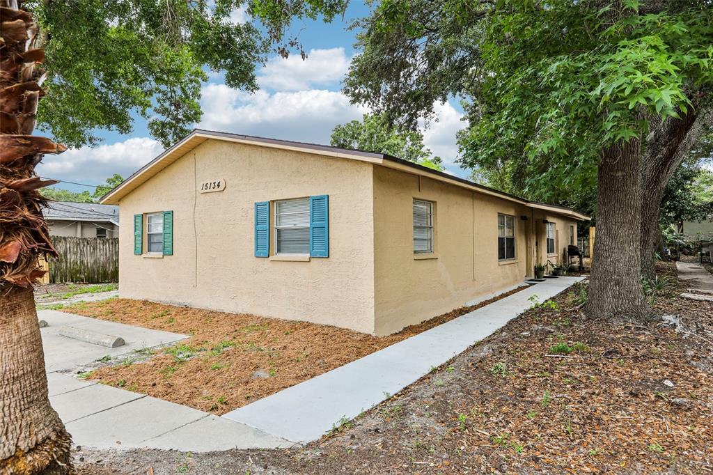 A single-story tan house with blue shutters sits on a lot with trees and concrete — 15134 VERONA AVENUE, CLEARWATER, FL