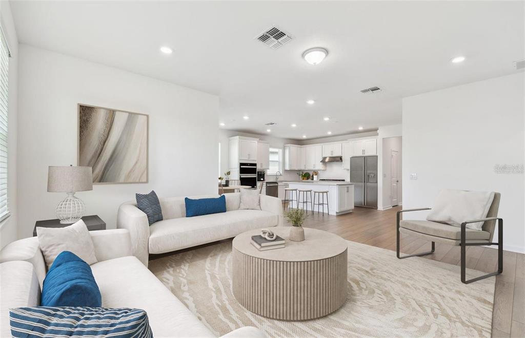 A bright open-concept living room features white sofas, a round coffee table, and a kitchen — 6741 MICHAEL FAVOR WAY, WESLEY CHAPEL, FL