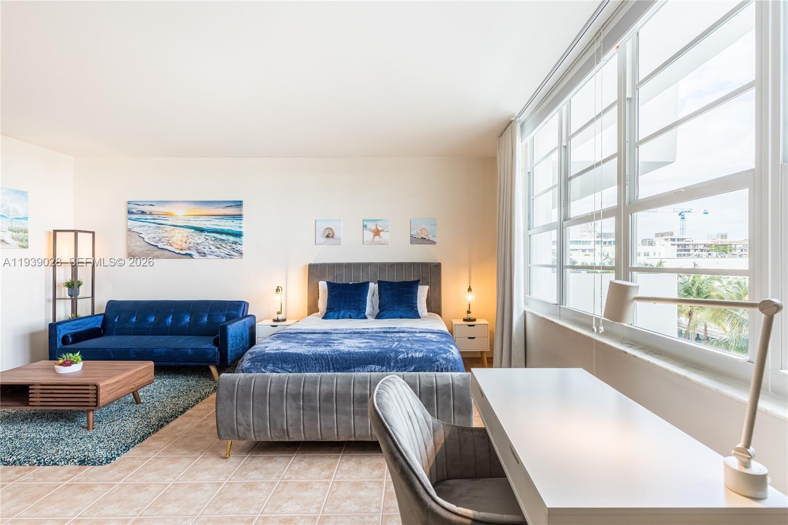 A bedroom features a bed, blue sofa, coffee table, desk, and large windows with views — 100 Lincoln Rd # 431, Miami Beach FL 33139, MIAMI BEACH, FL