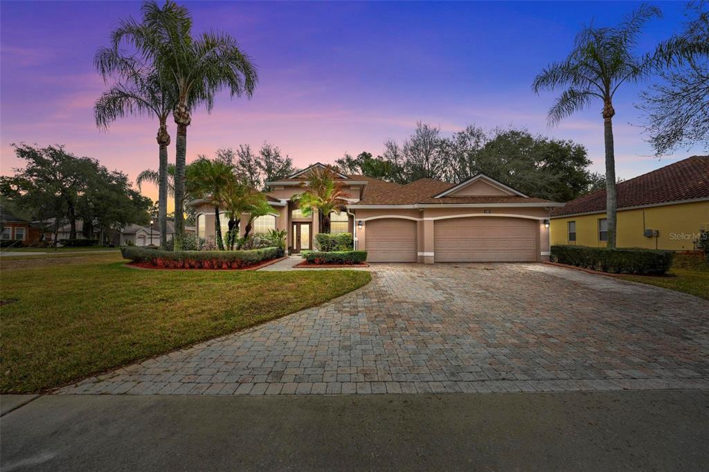 A single-story house with a paved driveway, three-car garage, and palm trees at sunset — 4385 SEA ROCK COURT, APOPKA, FL