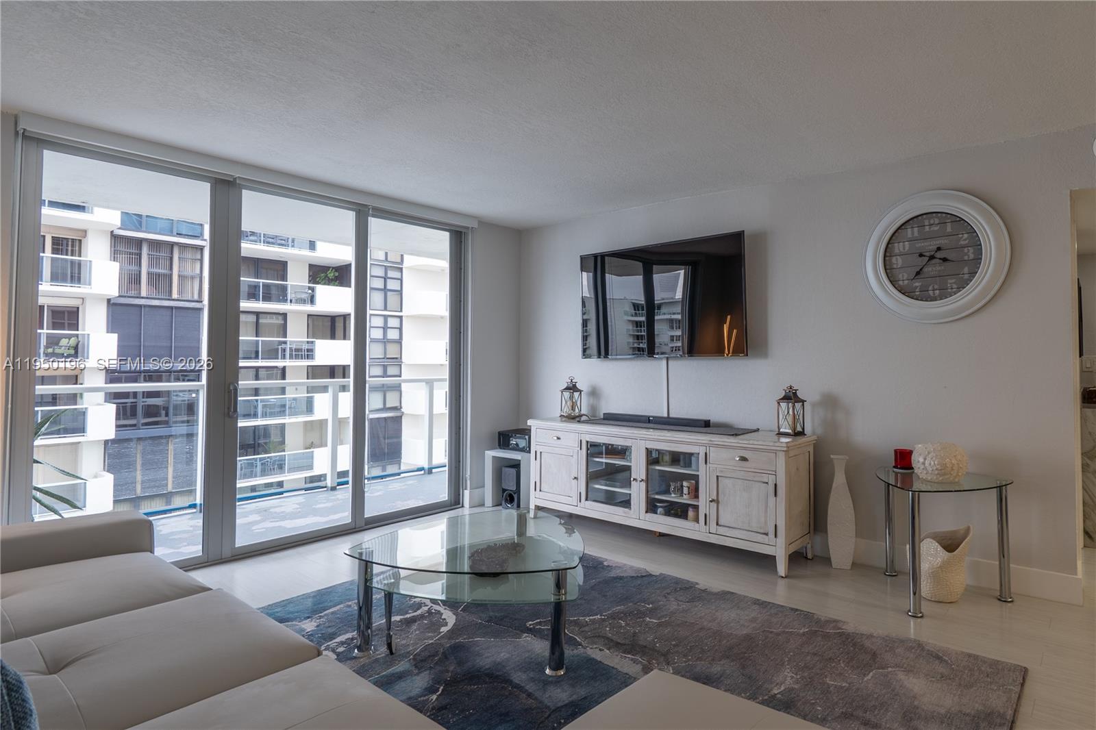 A living room features a glass coffee table, a white media console, and a balcony view — 5701 Collins Ave # 1417, Miami Beach FL 33140, MIAMI BEACH, FL