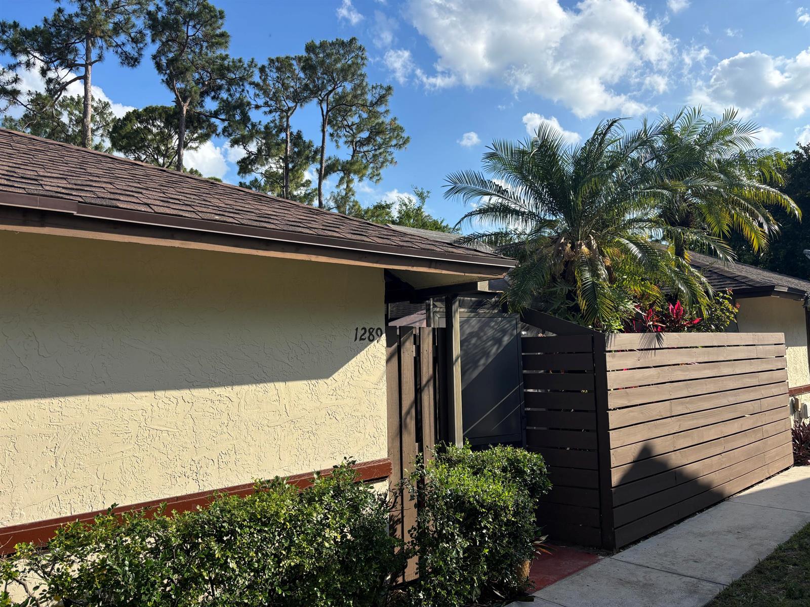A beige house exterior features a brown roof, wooden privacy fence, and a palm tree — 1289 Periwinkle Place, Wellington FL 33414, WELLINGTON, FL