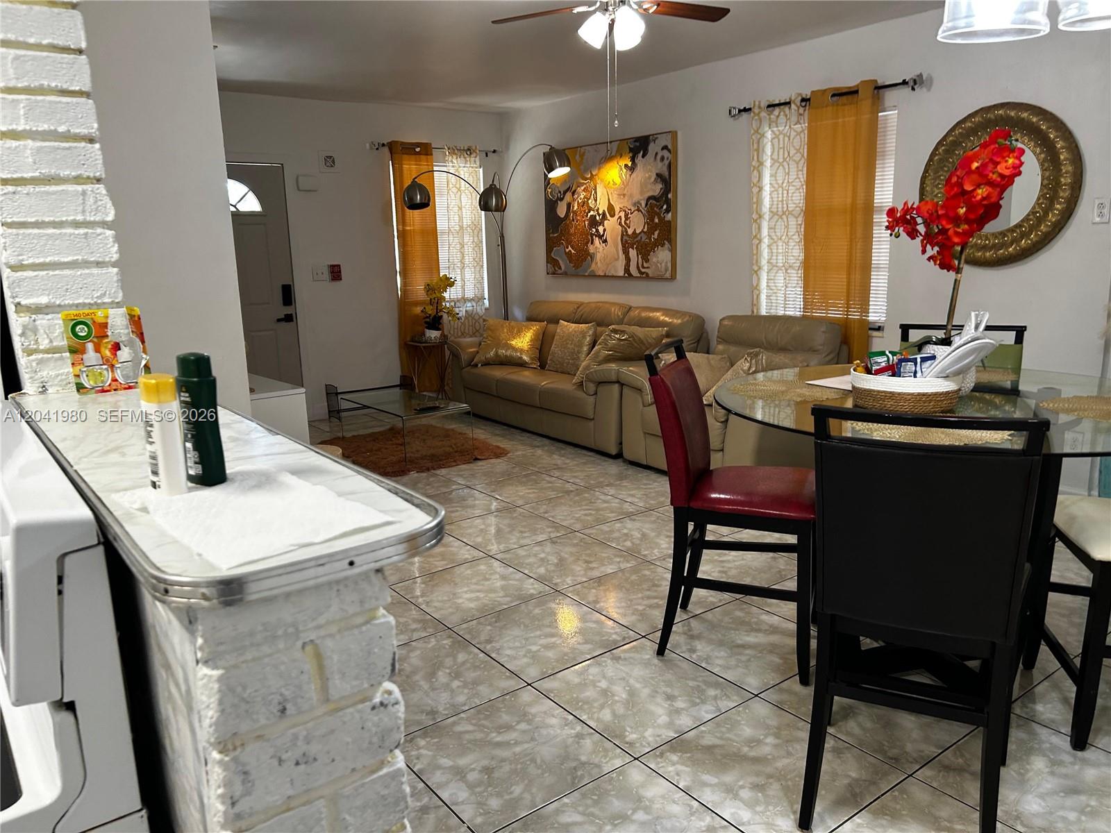 An open-concept living and dining area features tile flooring, a leather sofa, and gold accents — 295 NW 189th Ter, Miami Gardens FL 33169, MIAMI GARDENS, FL