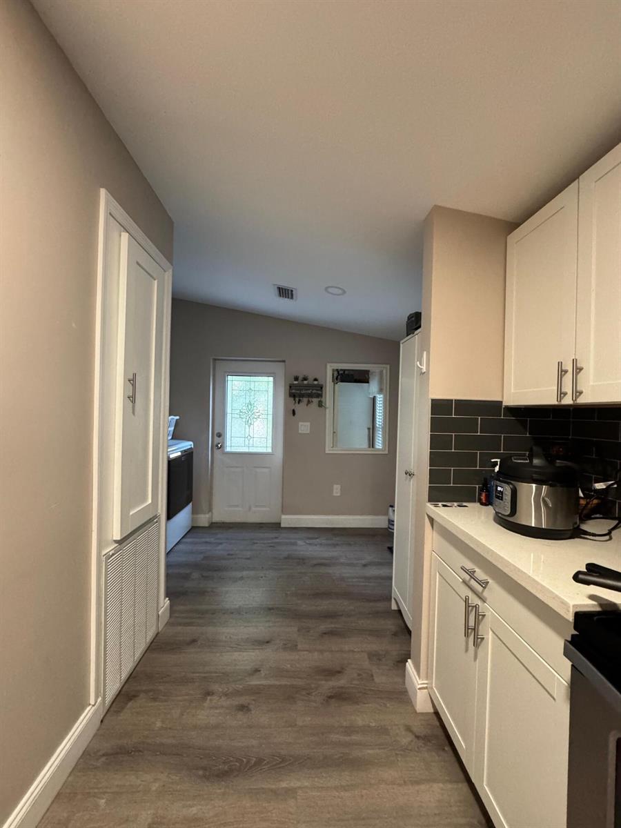 A kitchen with white cabinets, dark subway tile backsplash, and wood-look flooring leads to an entryway — 3899 Redman Parkway # 143, Boynton Beach FL 33436, BOYNTON BEACH, FL