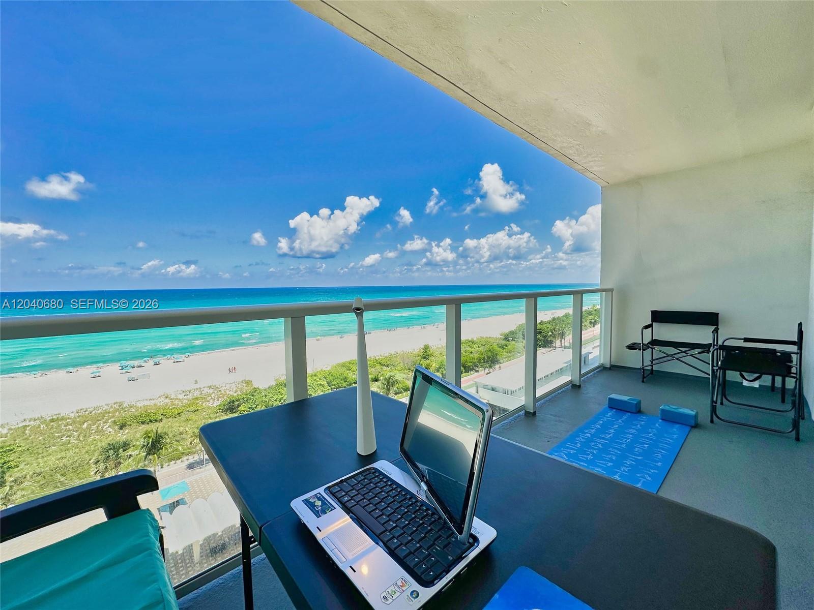 A balcony overlooks a sandy beach and the ocean, featuring a laptop on a table — 5701 Collins Ave, Miami Beach FL 33140, MIAMI BEACH, FL