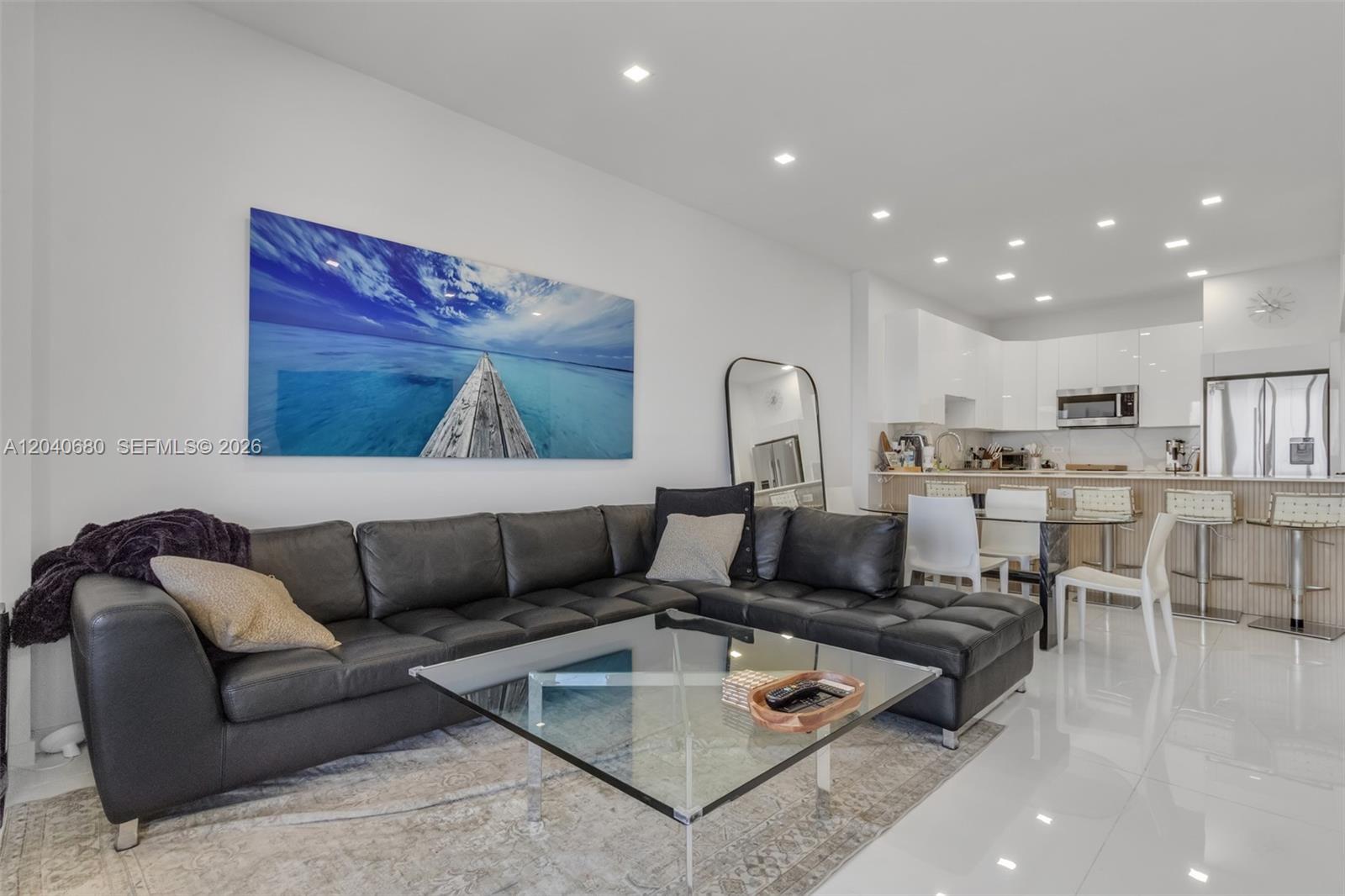 A modern living room features a black sectional sofa, glass coffee table, and open kitchen — 5701 Collins Ave, Miami Beach FL 33140, MIAMI BEACH, FL