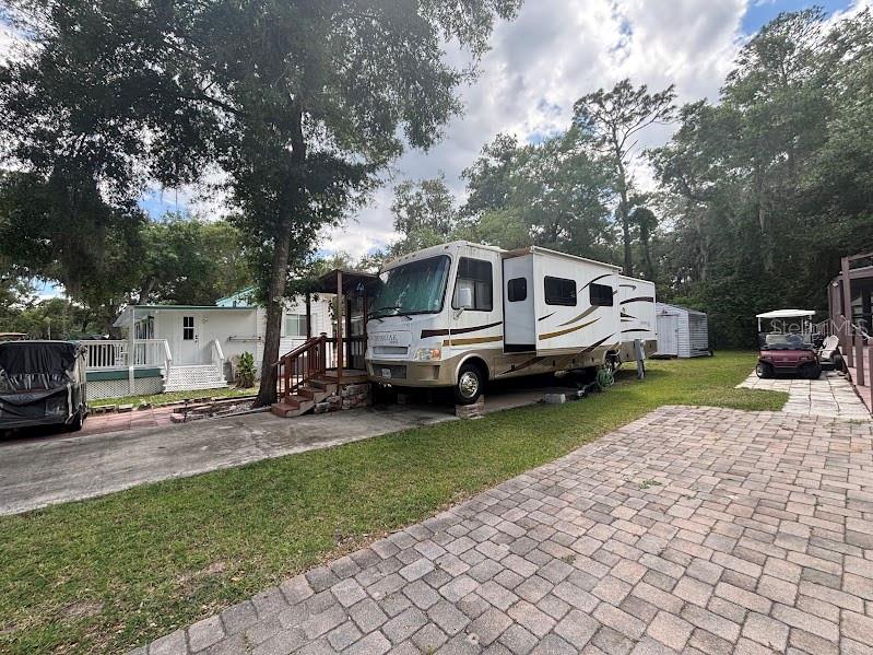 A motorhome is parked on a paved driveway next to a small house and trees — 14427 252ND COURT ROAD NE UNIT 138, FORT MC COY, FL