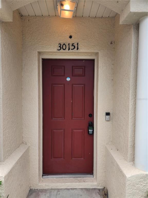 A red front door with a keypad lock is framed by beige stucco walls — 30151 BARNABY LANE, WESLEY CHAPEL, FL