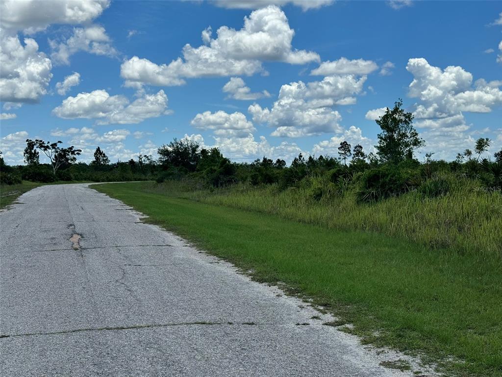 A paved road runs alongside a grassy, undeveloped lot under a bright, cloudy sky — Lot #2 MEADOWLARK BOULEVARD, INDIAN LAKE ESTATES, FL