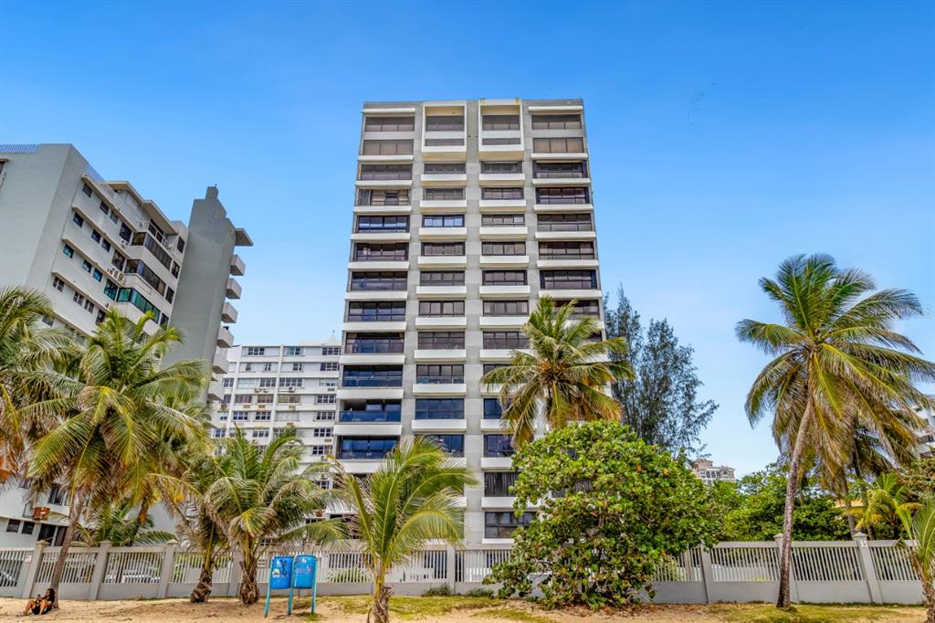 A tall, multi-story apartment building stands behind a fence and palm trees on a beach — 1507 ASHFORD AVE. UNIT 1002, SAN JUAN, PR