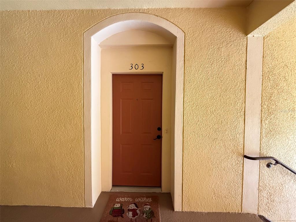 A brown apartment door labeled 303 features a decorative holiday doormat on a textured walkway — 6380 CONTESSA DRIVE UNIT 303, ORLANDO, FL