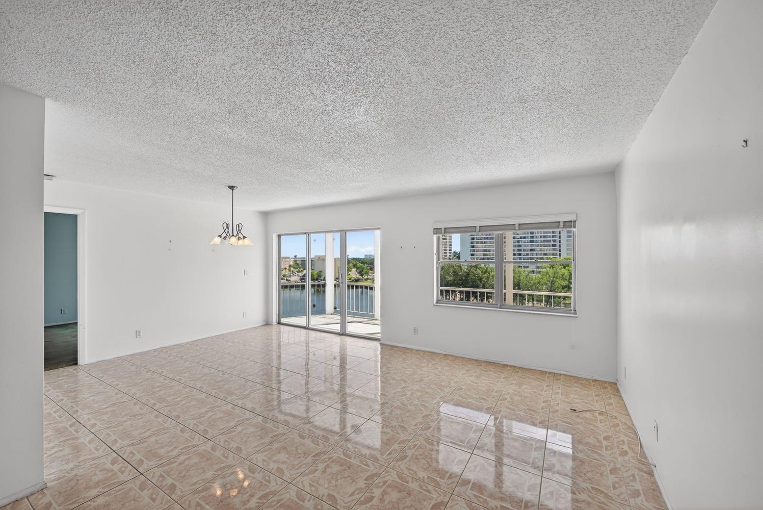 An empty living room with light-colored tile flooring, a popcorn ceiling, and a balcony view — 400 Diplomat Parkway # 612, Hallandale Beach FL 33009, HALLANDALE BEACH, FL