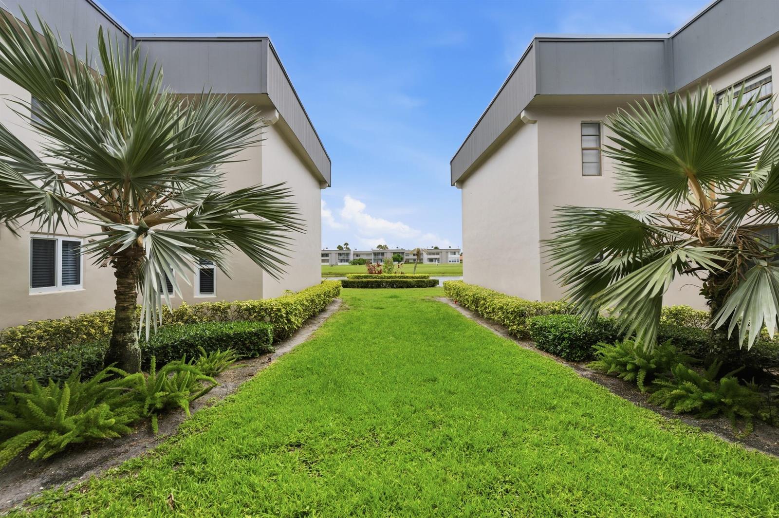 A grassy path between two apartment buildings leads toward a small pond and distant structures — 53 Piedmont B, Delray Beach FL 33484, DELRAY BEACH, FL
