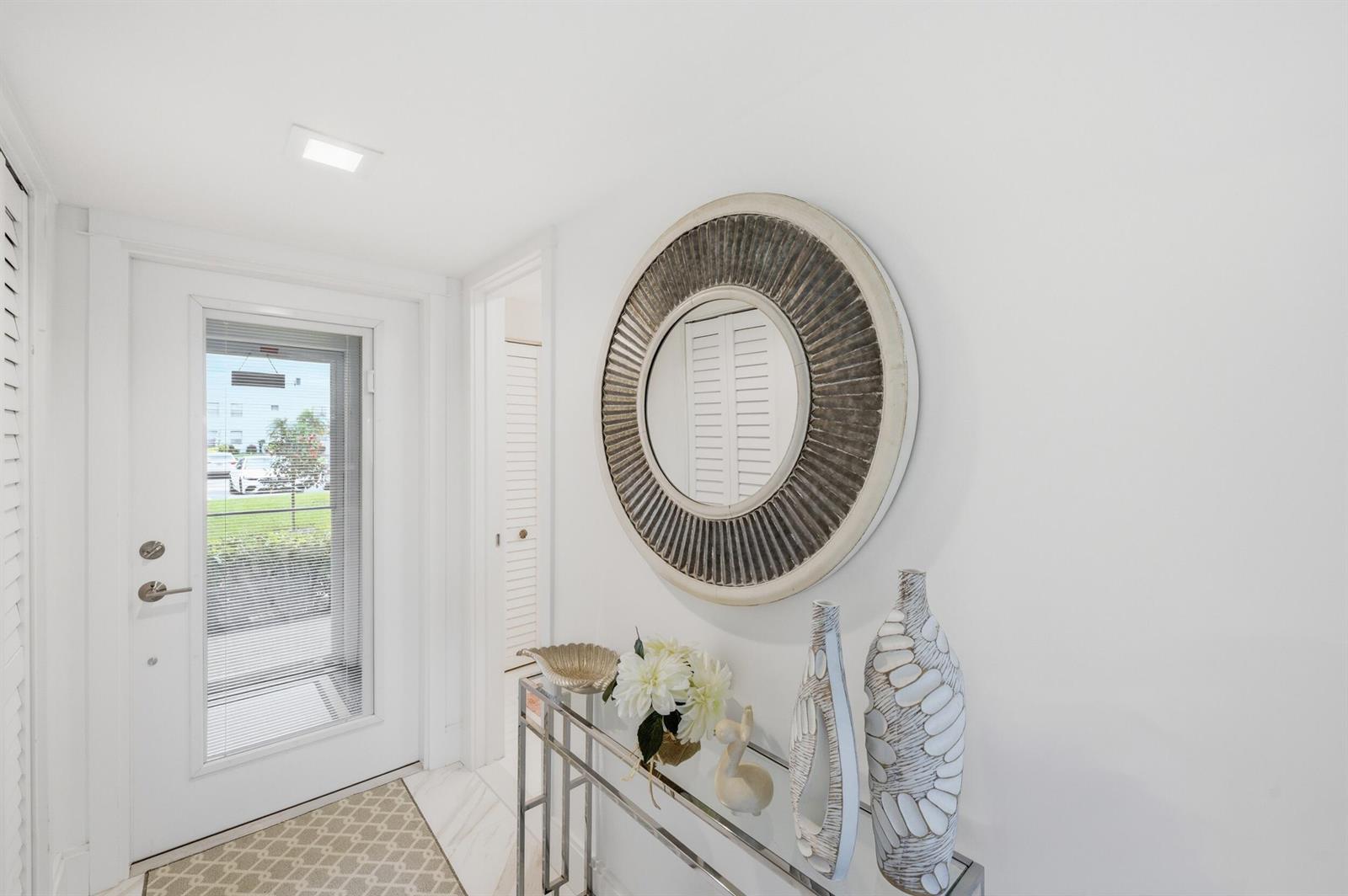 A bright entryway features a glass console table, a round mirror, and a white door — 53 Piedmont B, Delray Beach FL 33484, DELRAY BEACH, FL