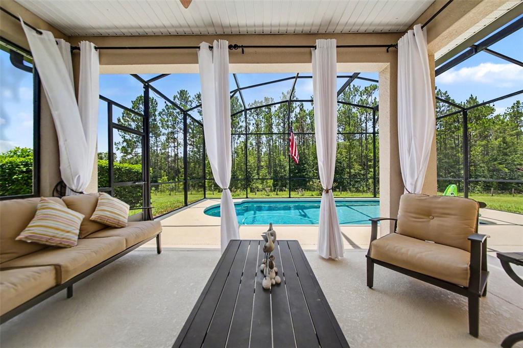 A covered patio features a seating area, white curtains, and a screened-in pool overlooking trees — 11200 BUCKHILL LANE, CLERMONT, FL