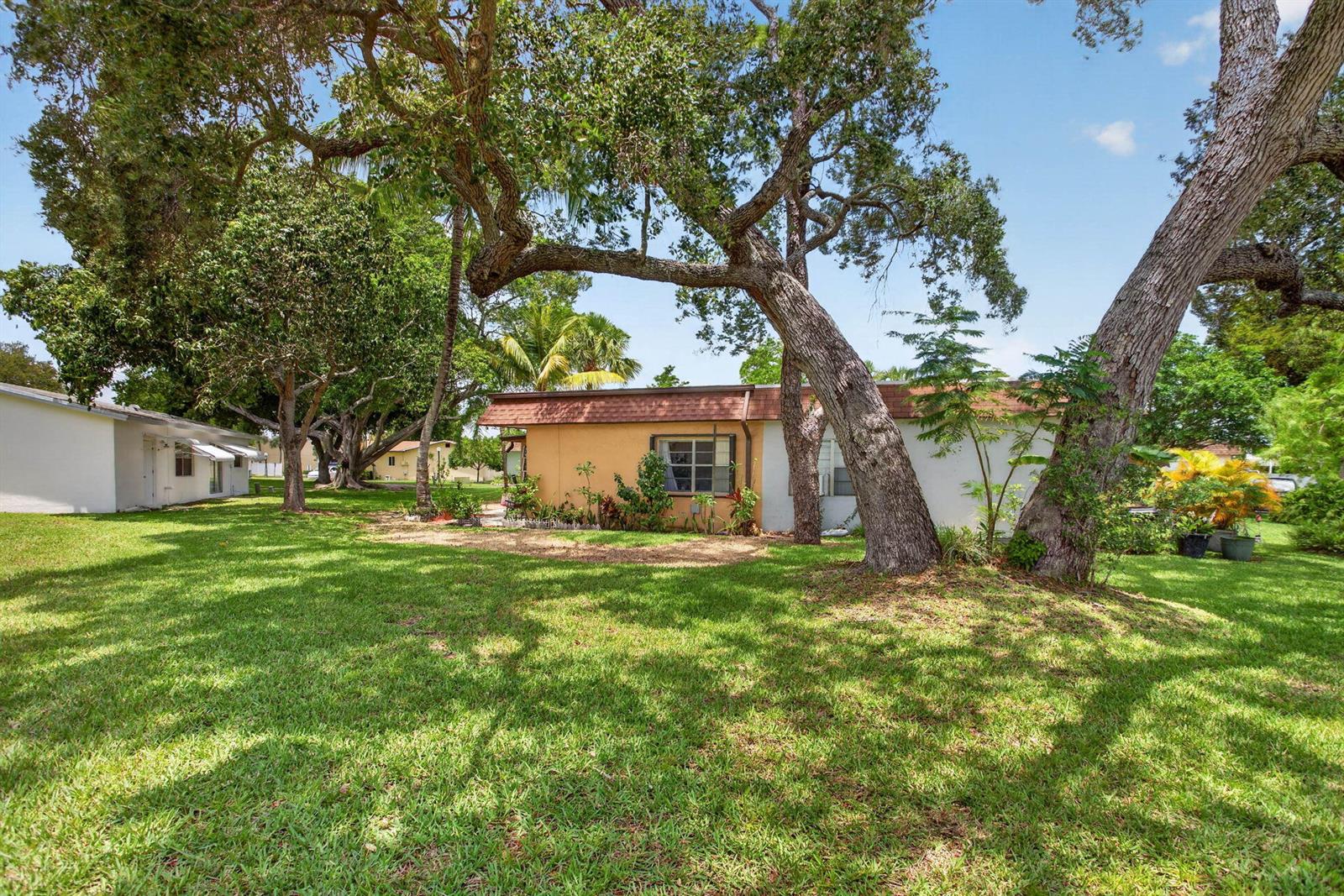 A single-story house with a tan exterior sits on a grassy lawn under large trees — 2002 SW Natura Boulevard # B, Deerfield Beach FL 33441, DEERFIELD BEACH, FL