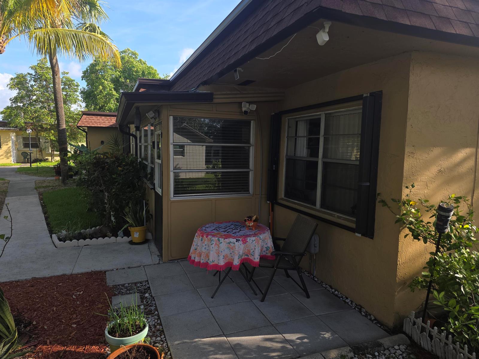 A patio with a small table and chair sits outside a tan house with windows — 2002 SW Natura Boulevard # B, Deerfield Beach FL 33441, DEERFIELD BEACH, FL