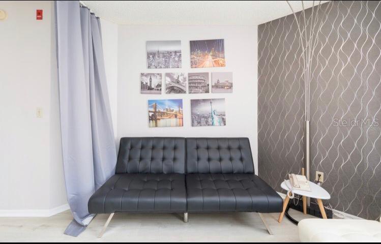 A living room features a black tufted futon, wall art, and a patterned accent wall — 6337 PARC CORNICHE DRIVE UNIT 2107, ORLANDO, FL