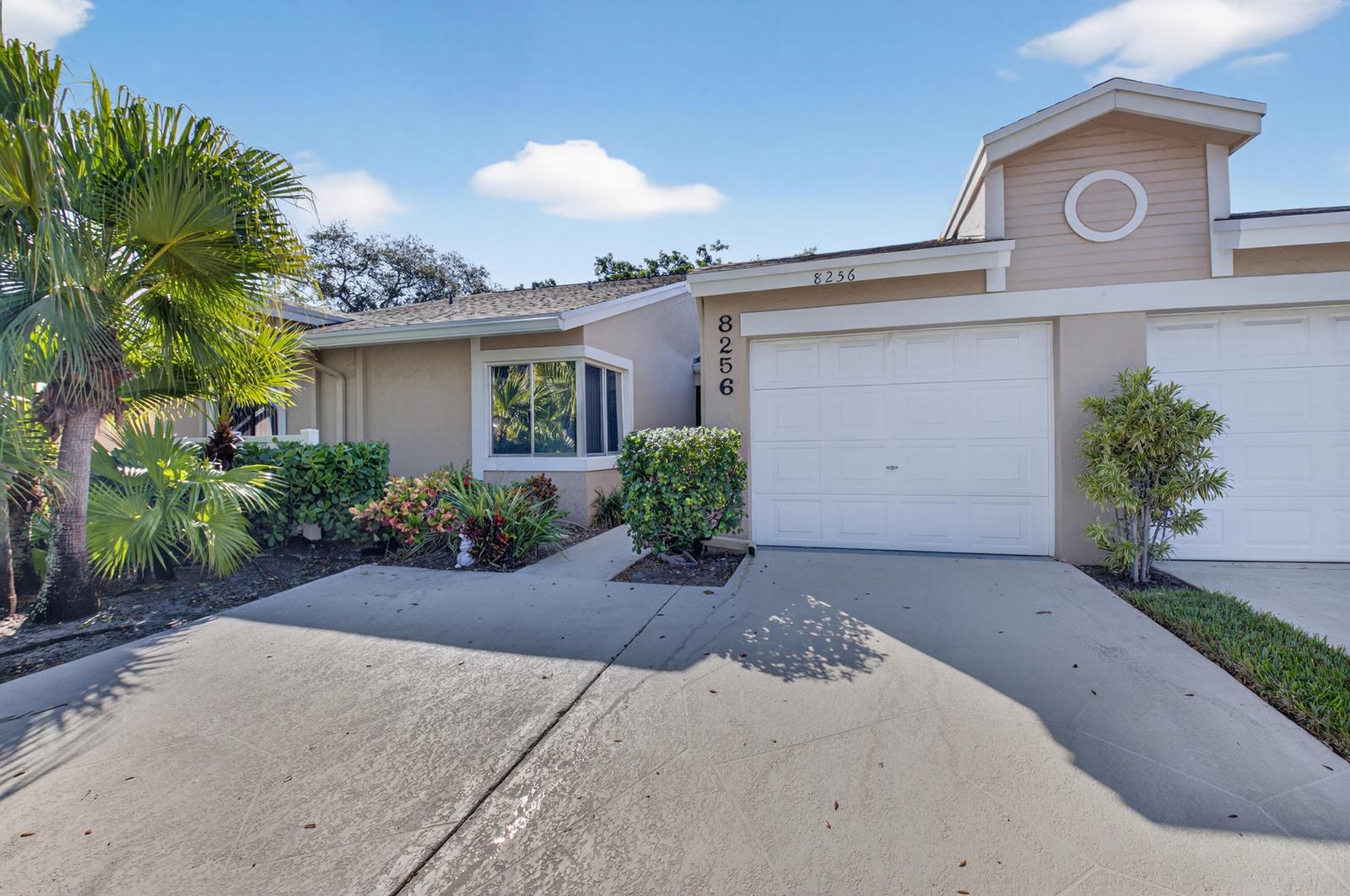 A tan single-story house features a white garage door, a concrete driveway, and palm trees — 8256 Summerbreeze Lane, Boca Raton FL 33496, BOCA RATON, FL
