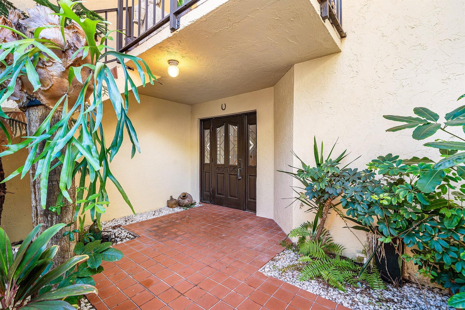An exterior entryway features a tiled patio, a dark wood front door, and lush plants — 16 Via Lago, Boynton Beach FL 33435, BOYNTON BEACH, FL