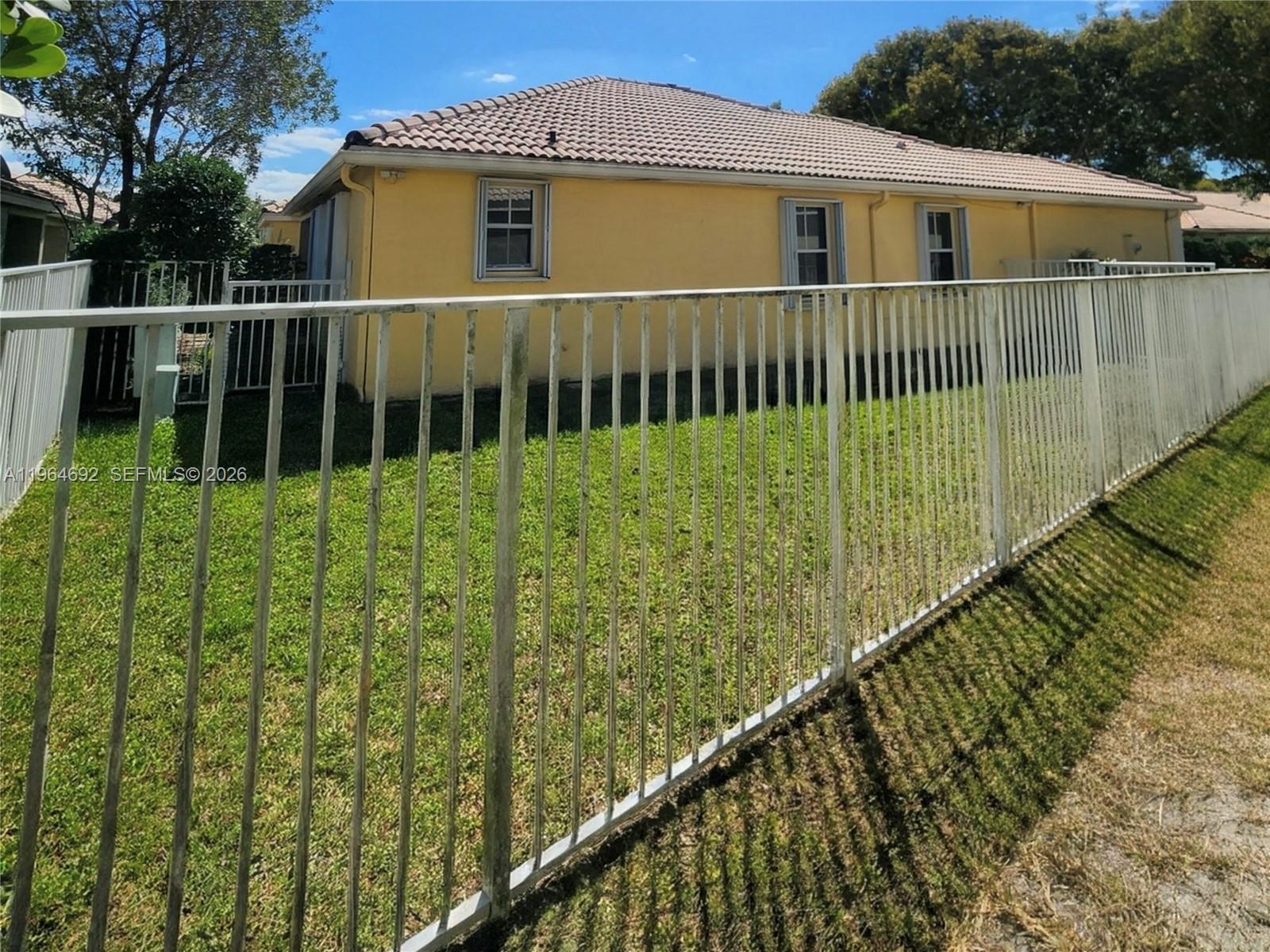 A yellow house with a tile roof is viewed from behind a white metal fence — 4309 Pine Ridge Ct, Weston FL 33331, WESTON, FL