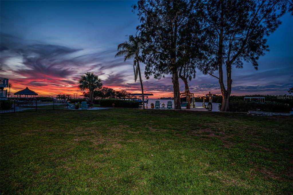 A grassy backyard overlooks a waterfront dock and palm trees during a vibrant sunset — 1626 SEA BREEZE DRIVE, TARPON SPRINGS, FL