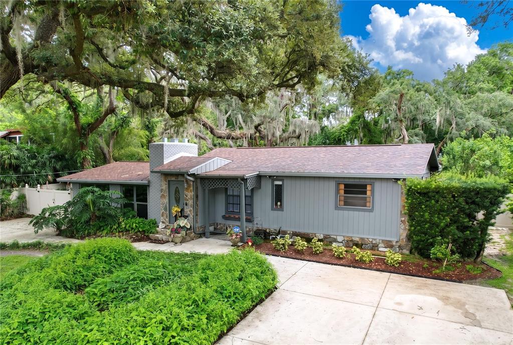 A single-story gray house with a stone chimney and a concrete driveway under large trees — 11805 N 51ST STREET, TEMPLE TERRACE, FL